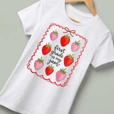 Whimsical illustration of strawberries with text 'first grade is my jam' in red and green — printed on a women's shirt