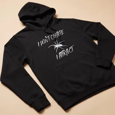 Minimalist spider design with bold text reading 'I Don’t Chase I Attract' in black and white. — printed on a unisex hoodie