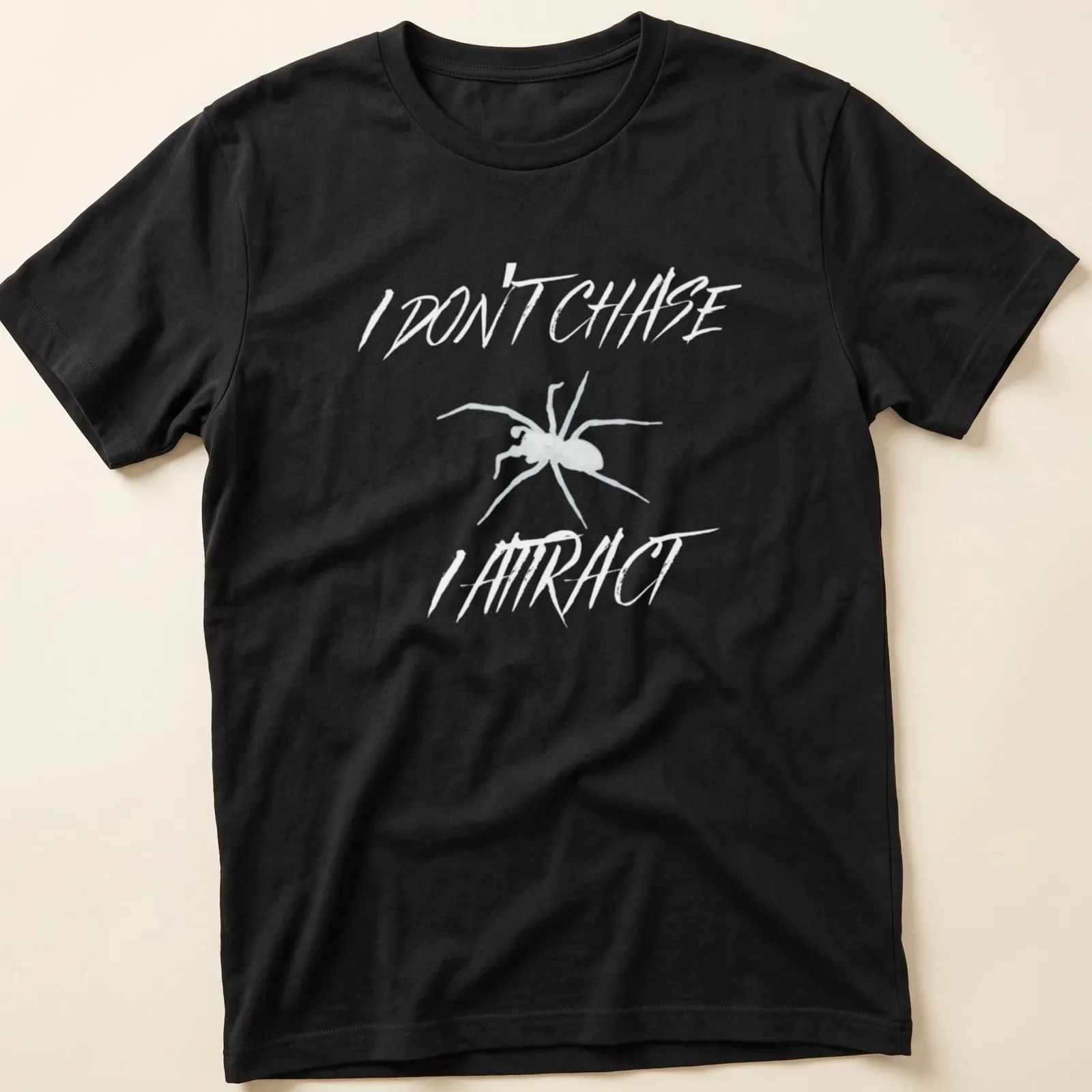 Minimalist spider design with bold text reading 'I Don’t Chase I Attract' in black and white. — printed on a unisex t-shirt