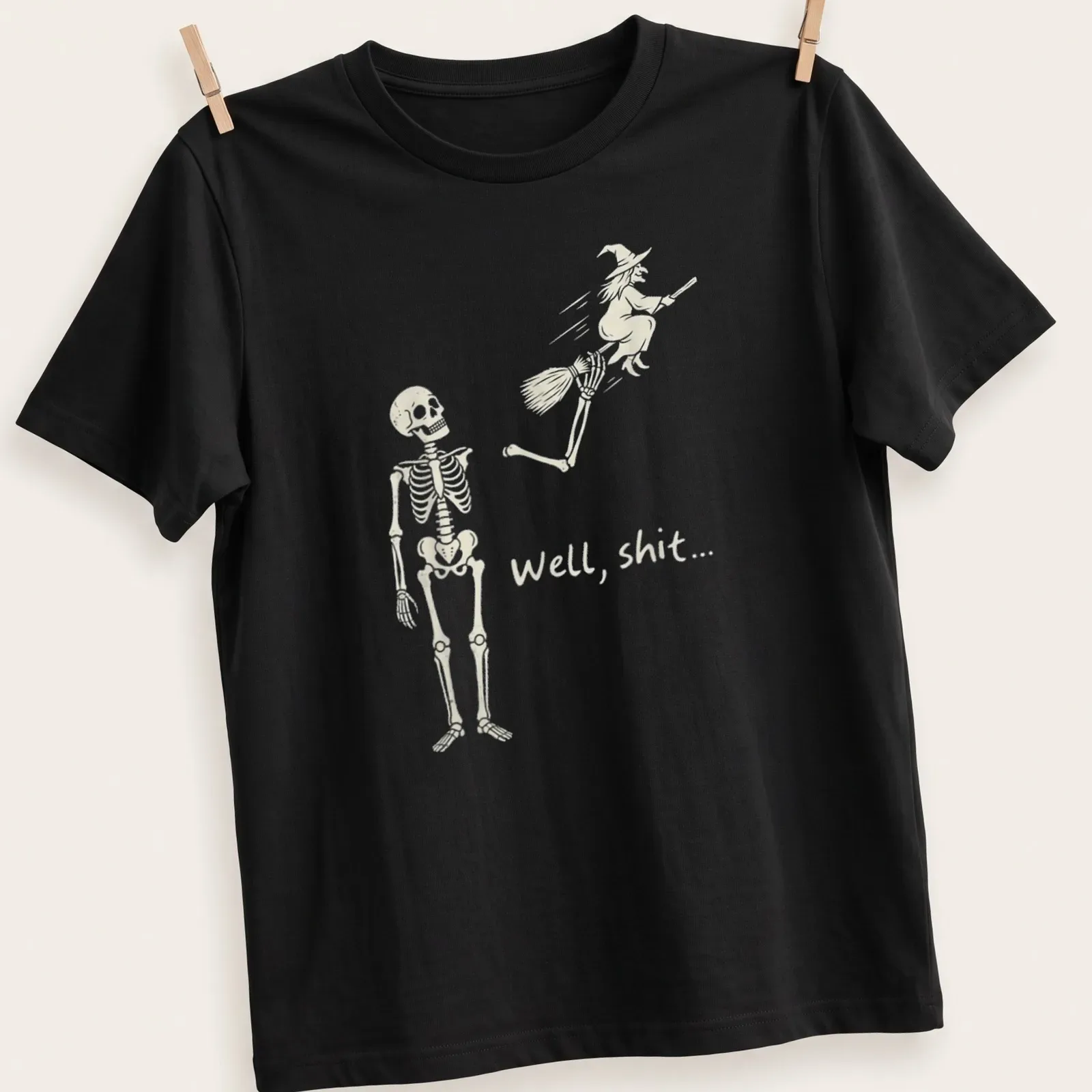 A humorous line-art print of a skeleton reacting to a flying witch with the text 'Well, shit...' — printed on a unisex t-shirt